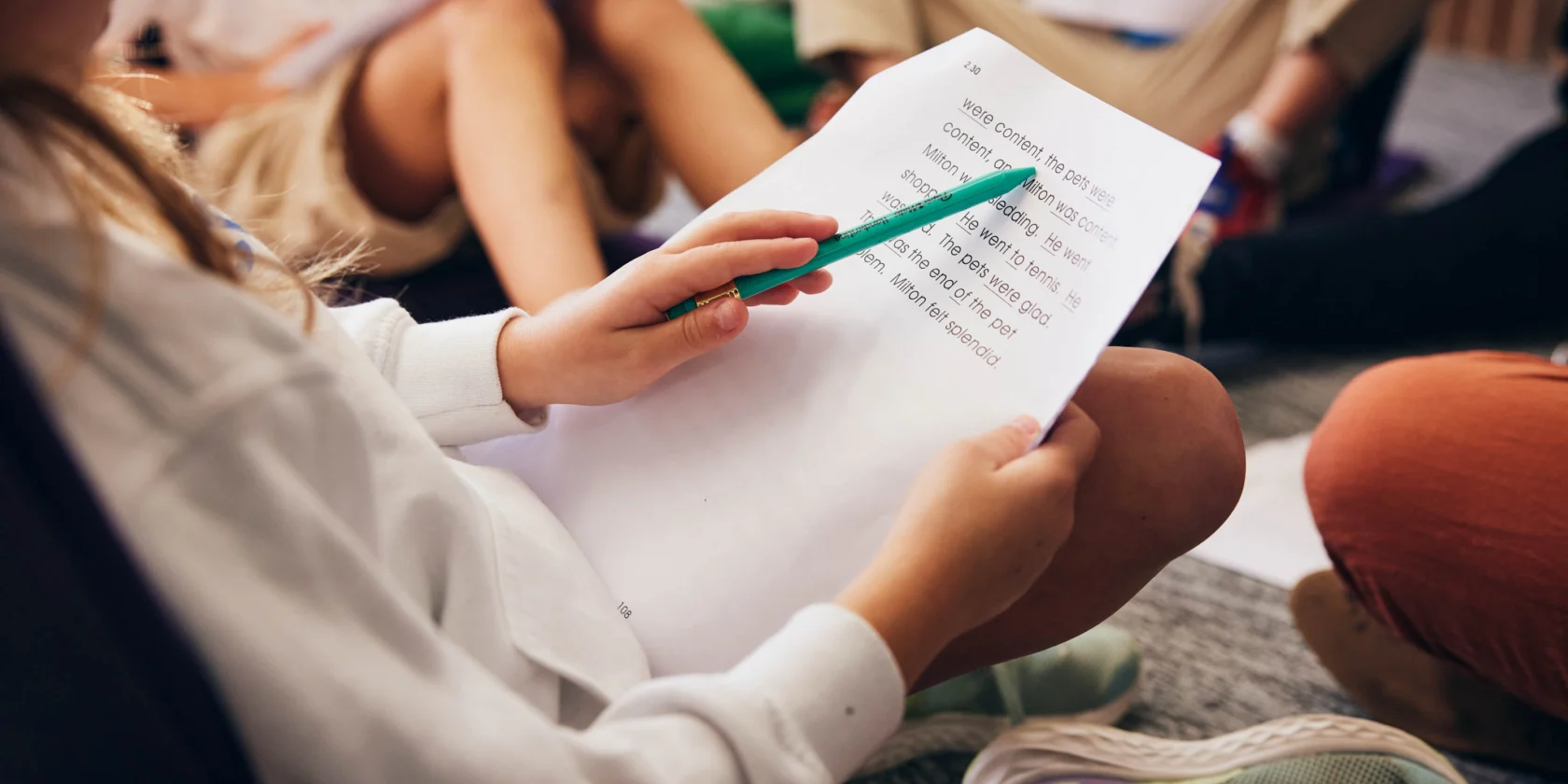 A person points at printed text on a sheet of paper with a green pencil, seated among others whose legs and papers are visible.