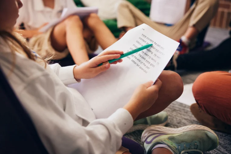 A person points at printed text on a sheet of paper with a green pencil, seated among others whose legs and papers are visible.