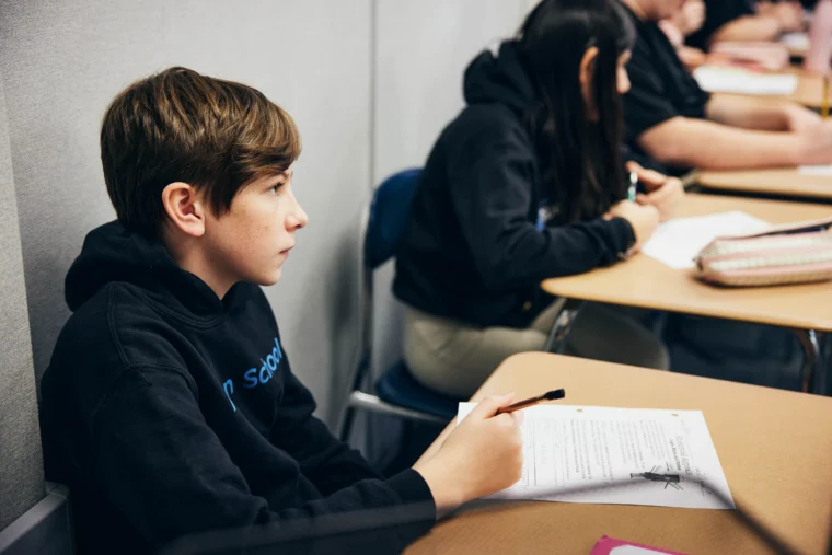 A student in a black hoodie sits at a desk holding a pen and looking forward, with papers and other students visible in the background.