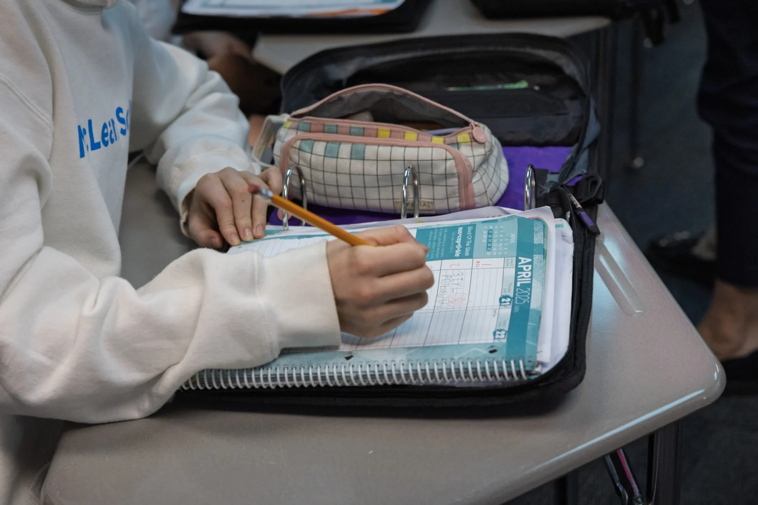 A student writes in a planner with a pencil at a desk, with a pencil case, binder, and notebook nearby.