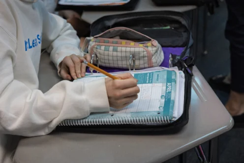 A student writes in a planner with a pencil at a desk, with a pencil case, binder, and notebook nearby.
