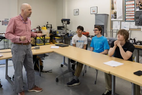 A teacher stands and speaks to three seated students in a classroom equipped with tools, equipment, and posters on the walls.