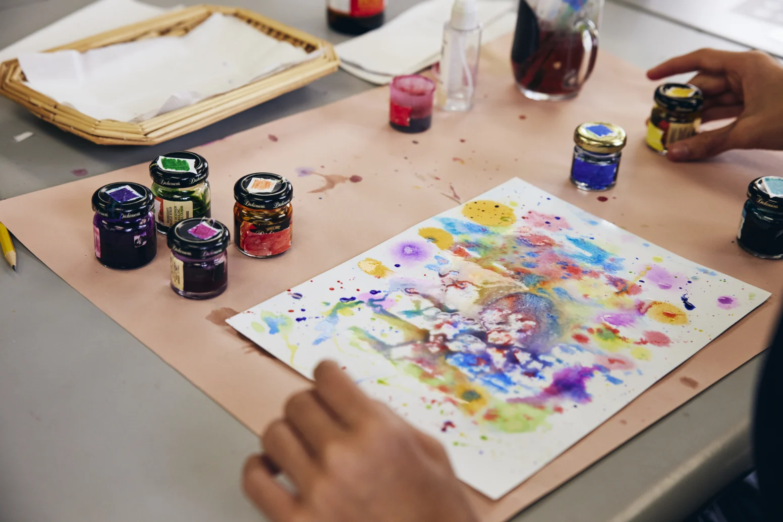 A person creates a colorful abstract painting with ink jars on a table, with art supplies and splattered paint visible.