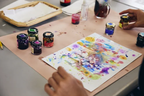 A person creates a colorful abstract painting with ink jars on a table, with art supplies and splattered paint visible.
