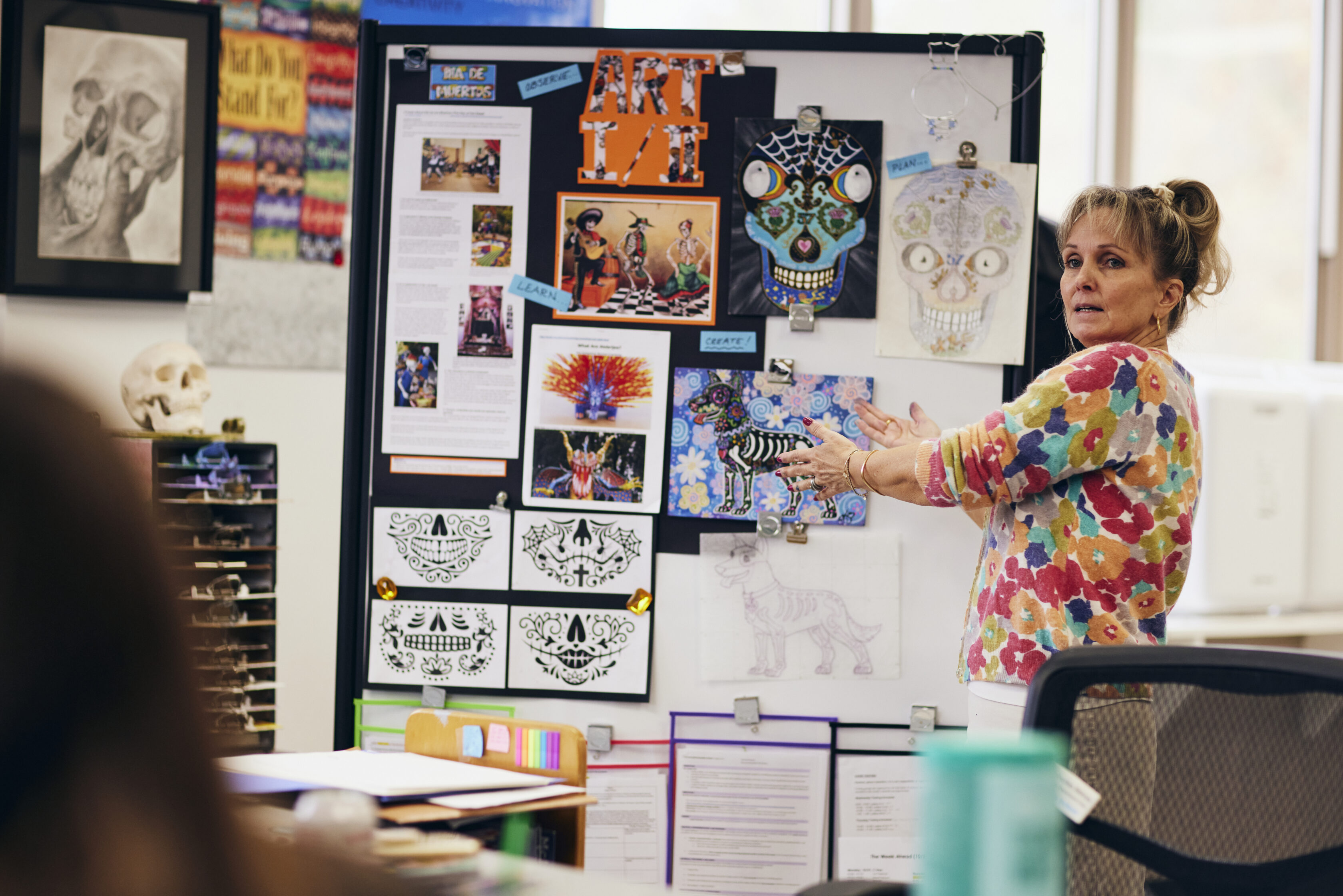 A woman stands and gestures towards an art display board covered with colorful drawings, photos, and printed materials in a classroom setting.