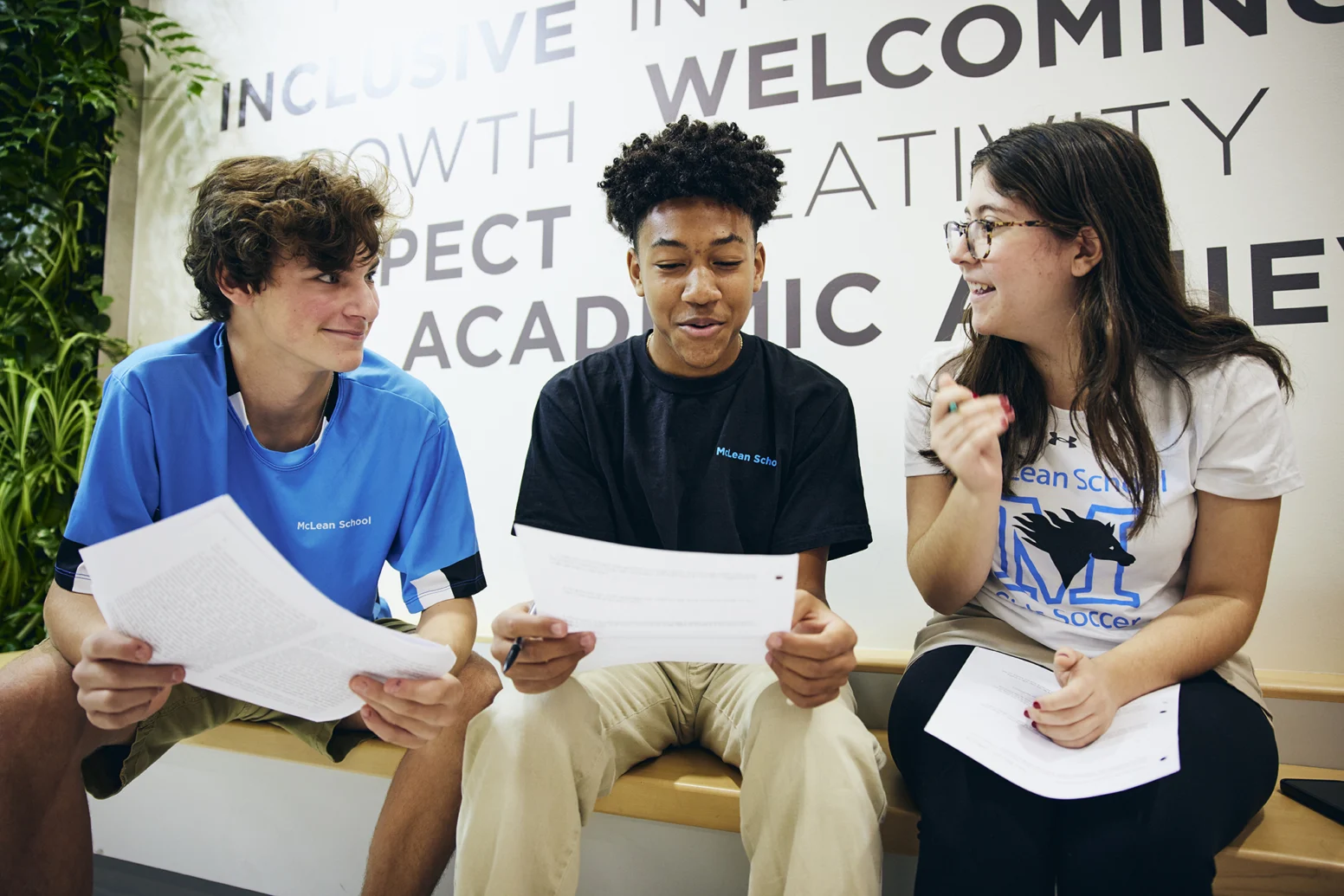 Three students sit on a bench indoors, holding papers and talking in front of a wall with large words like "welcoming" and "academic.