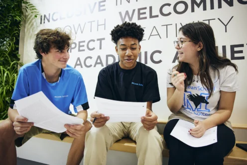 Three students sit on a bench indoors, holding papers and talking in front of a wall with large words like "welcoming" and "academic.