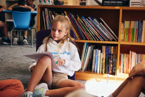 A girl sits on the floor in a classroom, holding papers and a pencil, with a bookshelf full of books behind her. Other students are partially visible nearby.