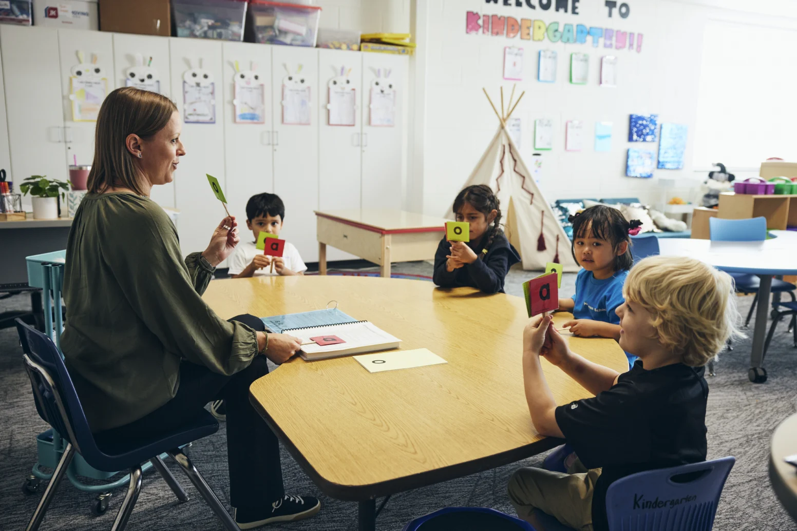 A teacher sits at a round table with four young children holding cards with letters in a classroom decorated with student artwork.