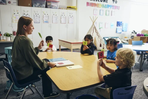 A teacher sits at a round table with four young children holding cards with letters in a classroom decorated with student artwork.