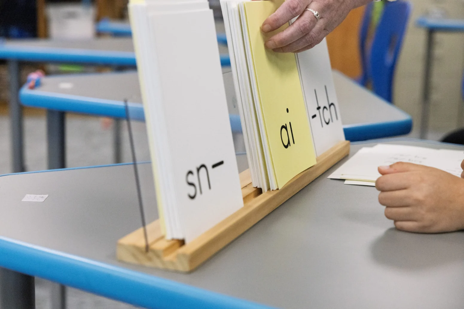 A hand arranges flashcards with the letter patterns "sn-", "ai", and "-tch" on a classroom desk, with another hand visible nearby.