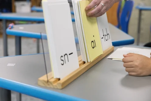 A hand arranges flashcards with the letter patterns "sn-", "ai", and "-tch" on a classroom desk, with another hand visible nearby.