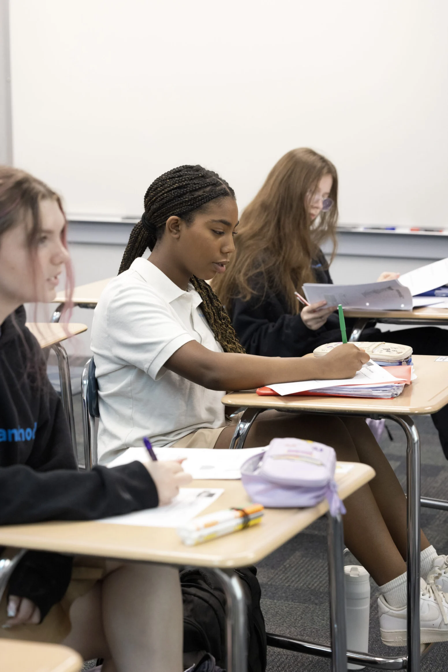Three students sit at desks in a classroom, writing in notebooks and focusing on their work.