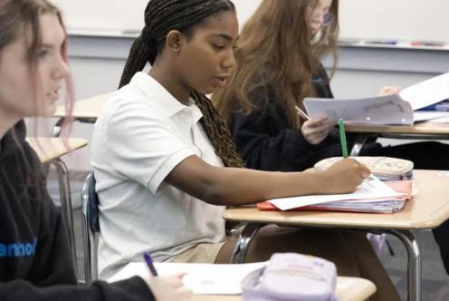 Three students sit at desks in a classroom, writing in notebooks and focusing on their work.