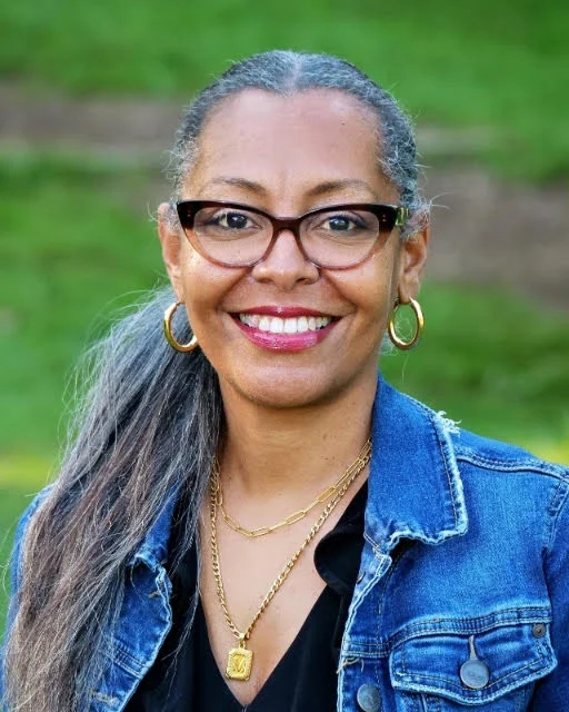 A woman with glasses and gray-streaked hair smiles outdoors, wearing a denim jacket, gold necklaces, and hoop earrings. Green grass is visible in the background.