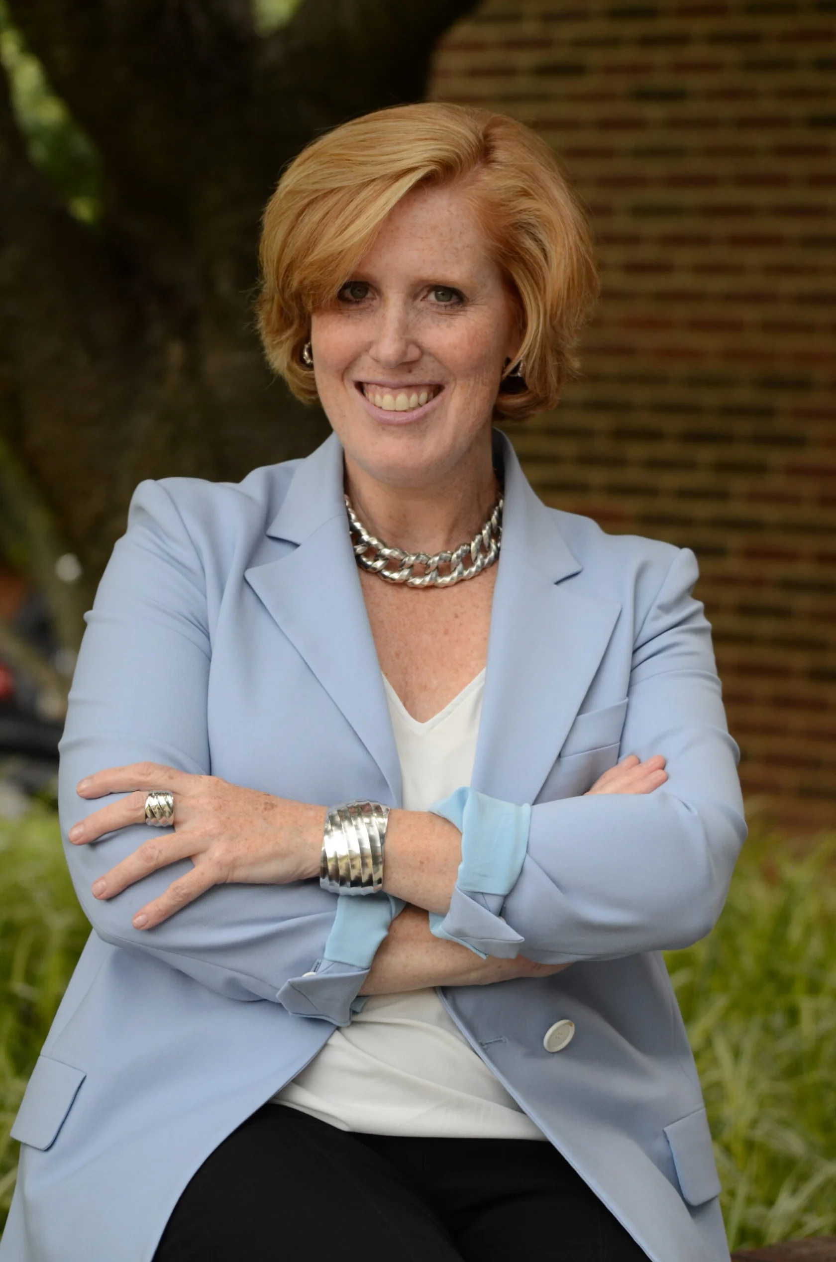 A woman with short red hair, wearing a light blue blazer, white top, and silver jewelry, sits outdoors with her arms crossed, smiling at the camera.