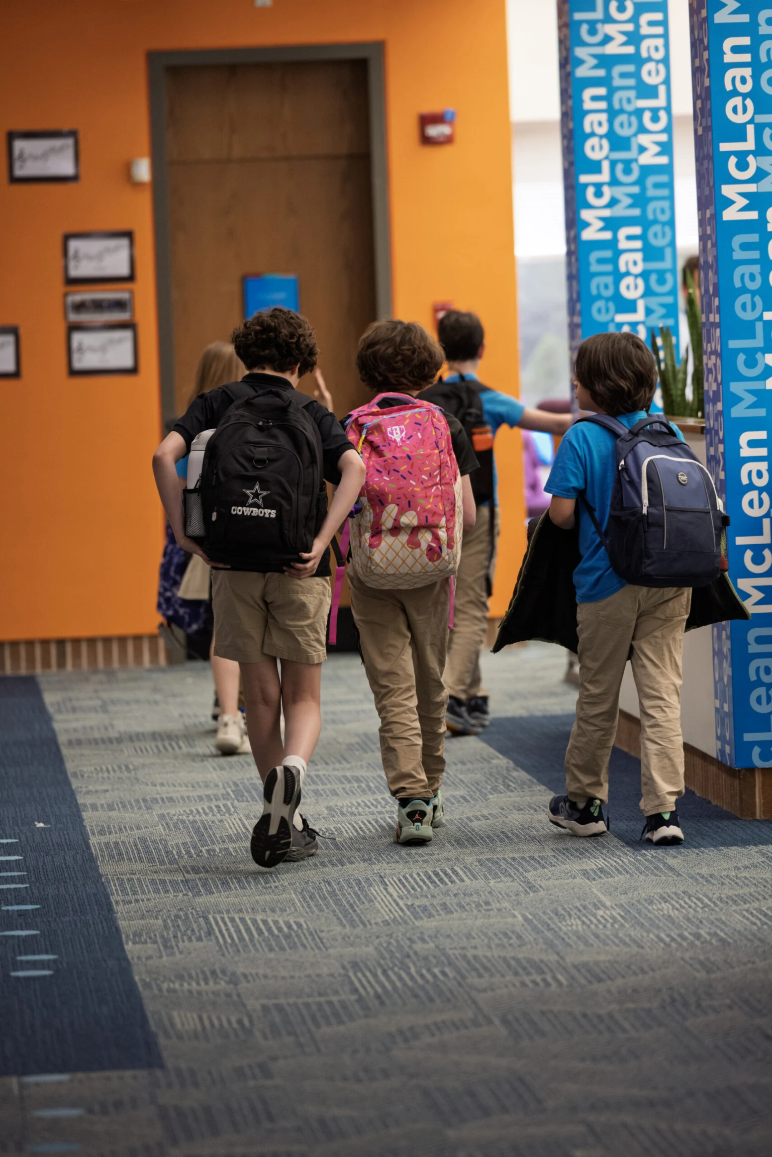 Four children with backpacks walk down a school hallway with orange and blue walls, heading toward a door.