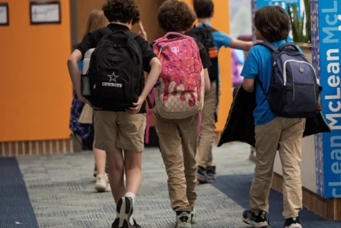 Four children with backpacks walk down a school hallway with orange and blue walls, heading toward a door.