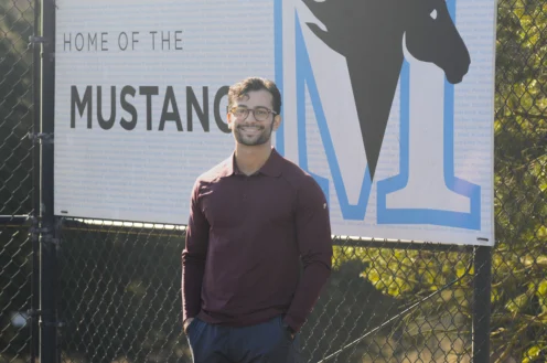 A man in a maroon shirt stands in front of a sign that reads "Home of the Mustangs" with a large horse graphic and the letter M.