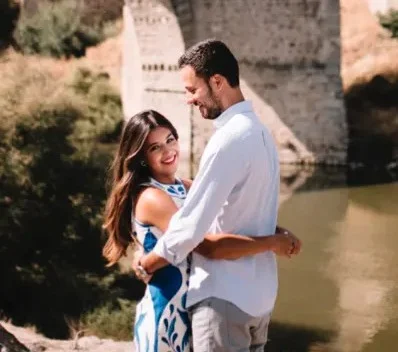 A couple stands embracing near a river, smiling at the camera, with a stone bridge and greenery in the background.