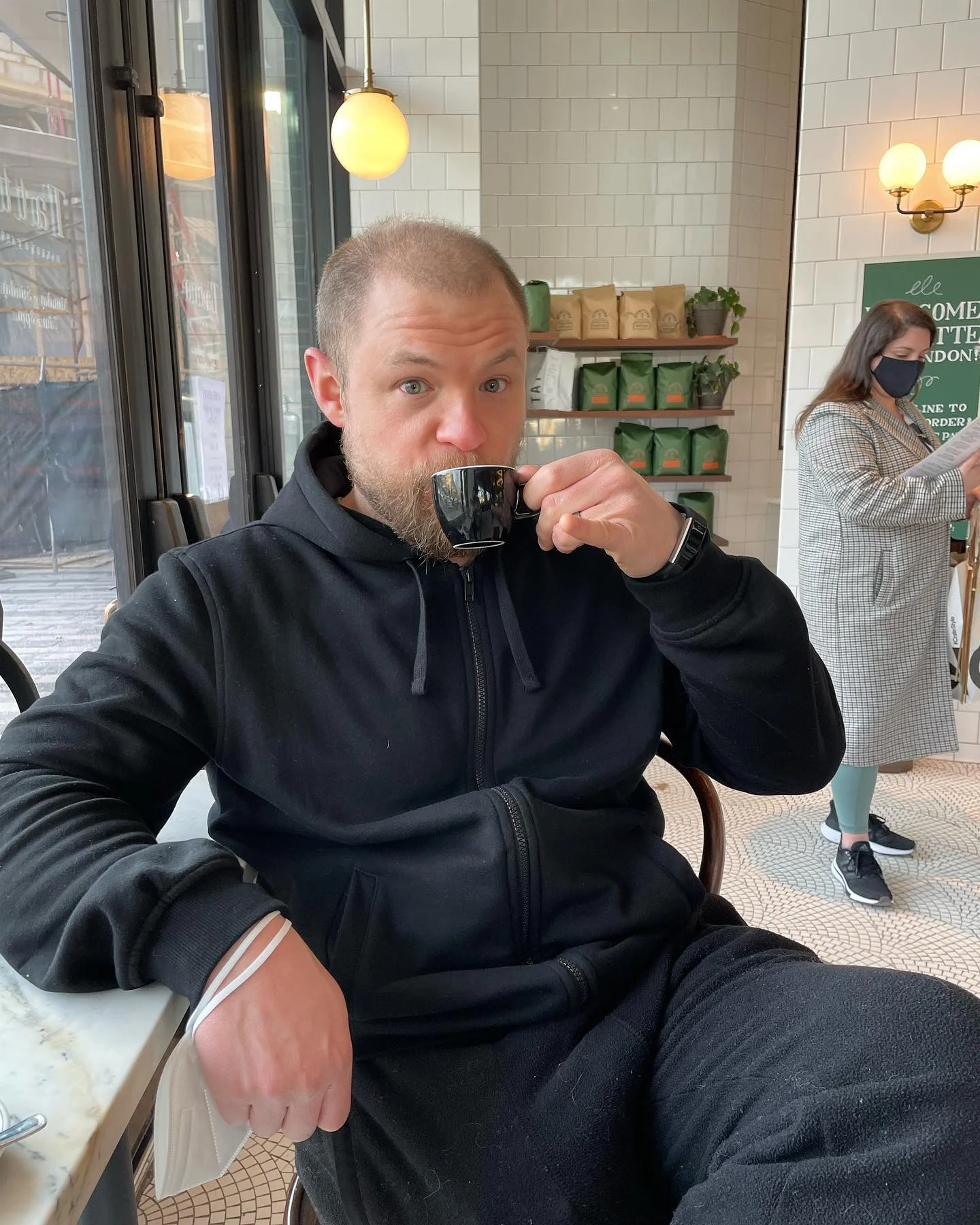 A man in a black hoodie sits at a café table, drinking from a small black cup. Behind him, a woman in a coat and mask uses her phone near shelves of packaged goods.