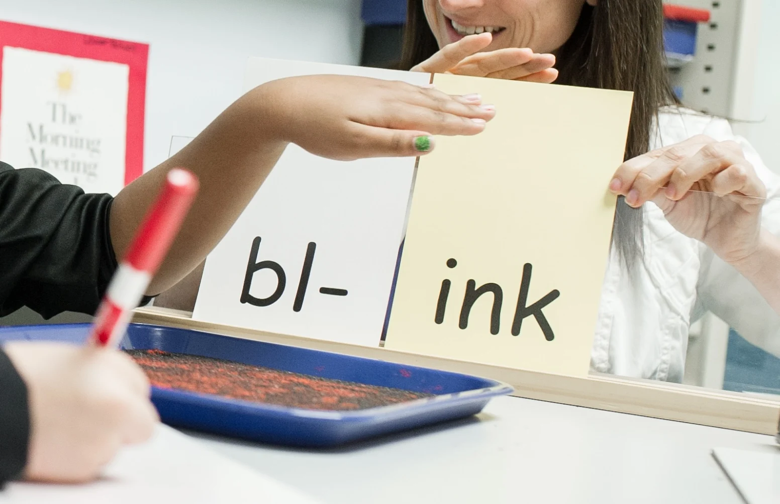 A child and adult practice reading the word "blink" using flashcards with "bl-" and "ink" written on them. A tray with red sand and a marker are on the table.