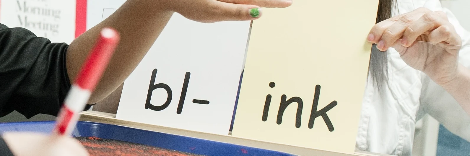 A child and adult practice reading the word "blink" using flashcards with "bl-" and "ink" written on them. A tray with red sand and a marker are on the table.