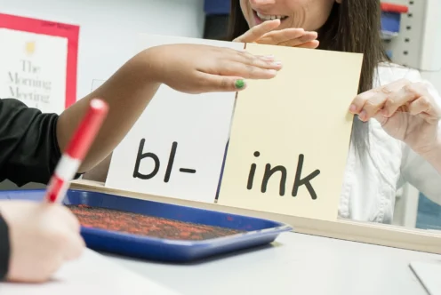 A child and adult practice reading the word "blink" using flashcards with "bl-" and "ink" written on them. A tray with red sand and a marker are on the table.
