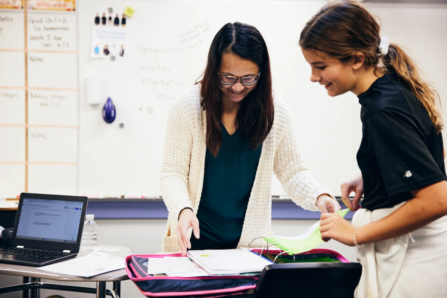 A teacher and a student look at papers together at a classroom desk, with a laptop and an open binder in front of them. A whiteboard with writing is visible in the background.
