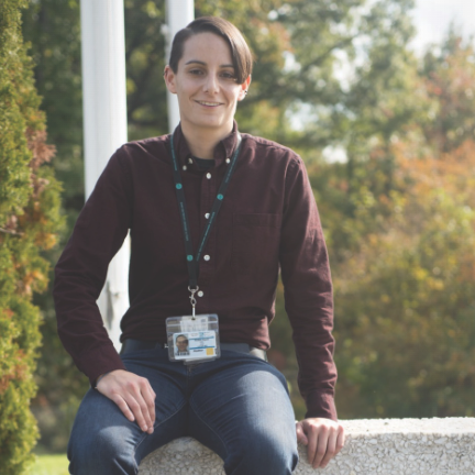 A person wearing a maroon shirt and ID badge sits on a stone bench outdoors with trees in the background.
