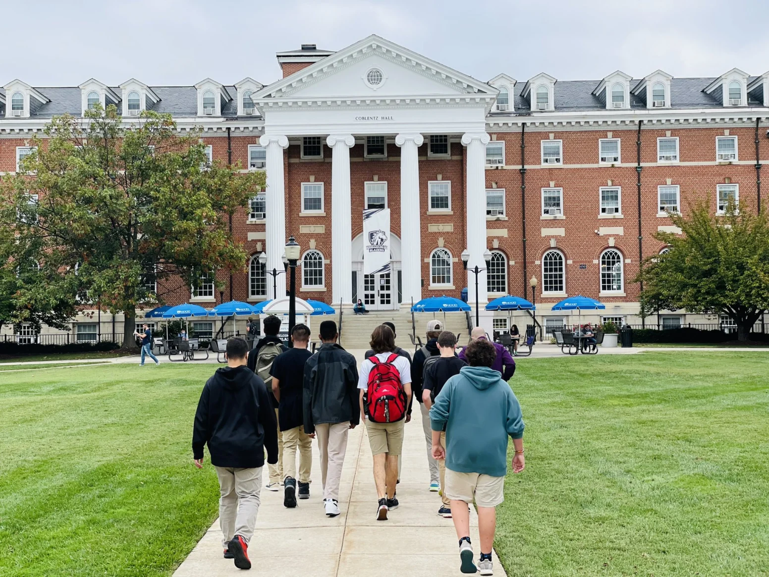 A group of students walks toward a large brick academic building with white columns and a flag out front on a cloudy day.