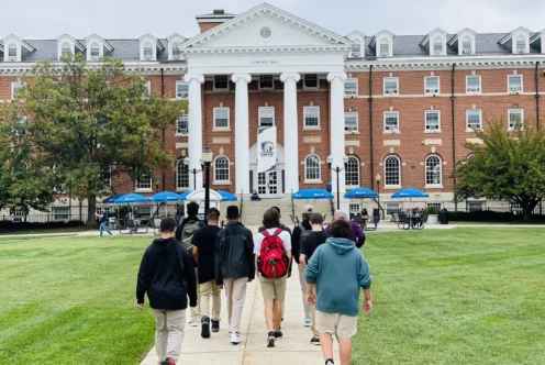 A group of students walks toward a large brick academic building with white columns and a flag out front on a cloudy day.