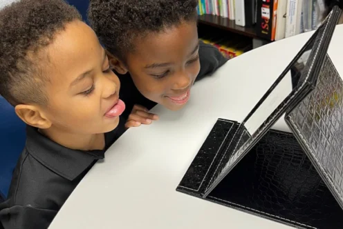 Two young students sit at a table, looking into a mirror and practicing tongue placement for speech sounds during a speech-language pathology session.