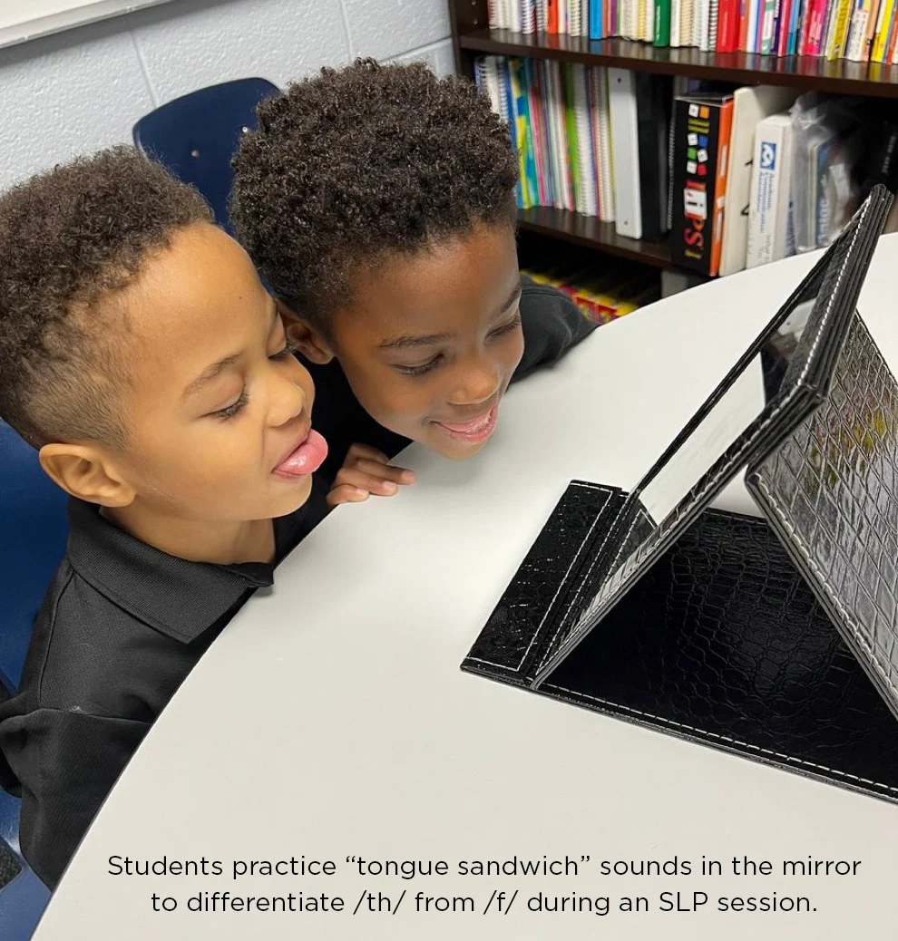 Two young students sit at a table, looking into a mirror and practicing tongue placement for speech sounds during a speech-language pathology session.