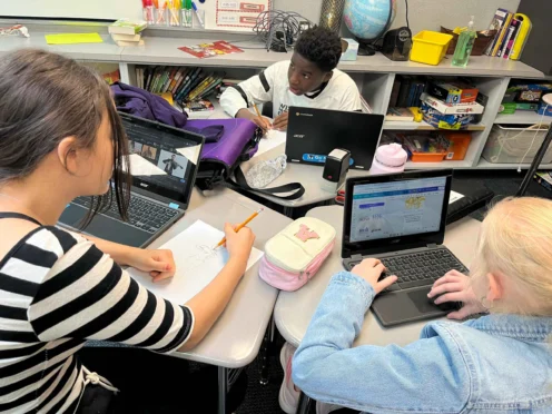 Three students sit at a table working on laptops and drawing, surrounded by school supplies and books in a classroom setting.