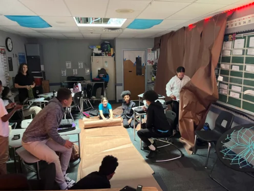 A group of students and a teacher work together in a classroom, spreading large brown sheets of paper on the floor and walls for a project.