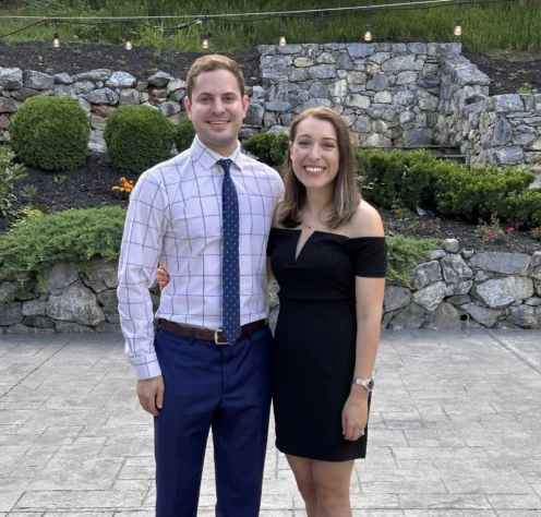 A man in a checkered shirt and tie stands next to a woman in a black off-the-shoulder dress; both are smiling outdoors in front of stone landscaping.