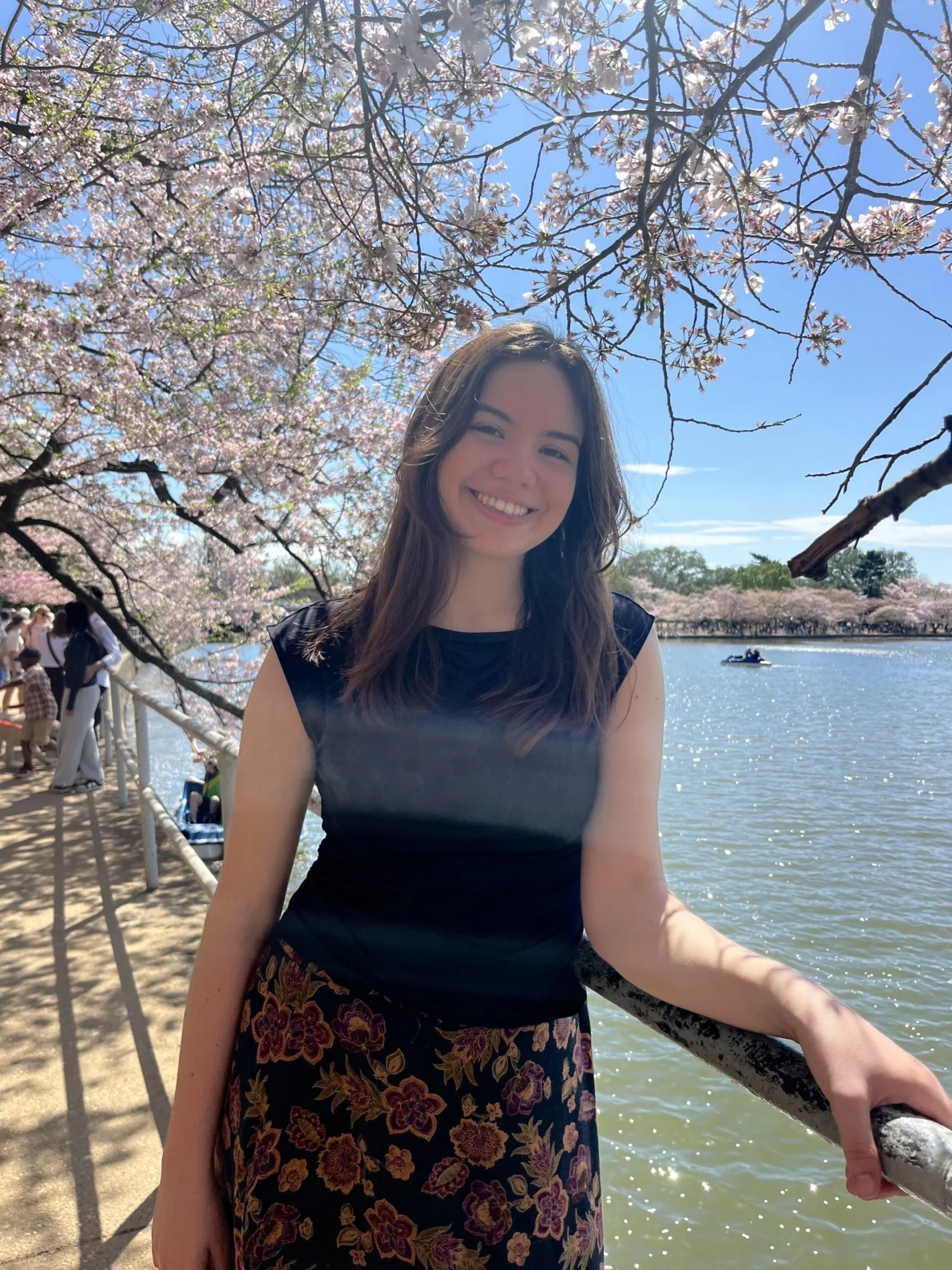A woman stands by a lakeside railing under blooming cherry blossom trees, smiling at the camera on a sunny day.