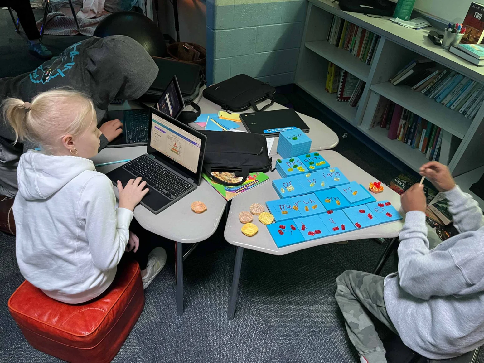 Three students work at small tables; two use laptops while one arranges game pieces on a board. Books and snacks are nearby.