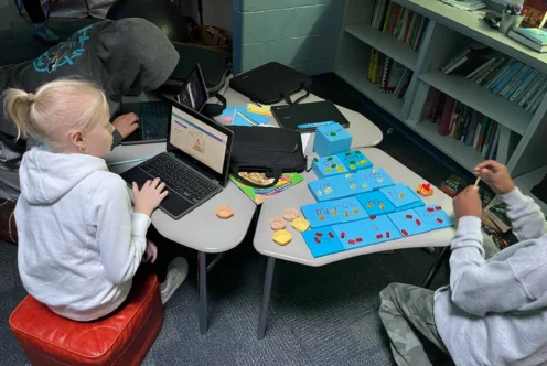 Three students work at small tables; two use laptops while one arranges game pieces on a board. Books and snacks are nearby.
