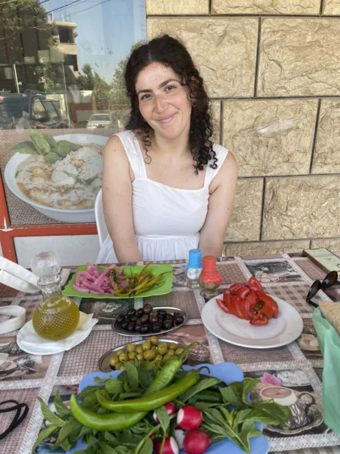 A woman in a white dress sits at an outdoor table with plates of vegetables, pickles, olives, and a bottle of olive oil in front of her.