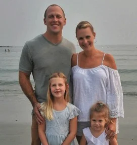 A man and woman stand together on a beach with two young girls in front of them, all smiling at the camera.
