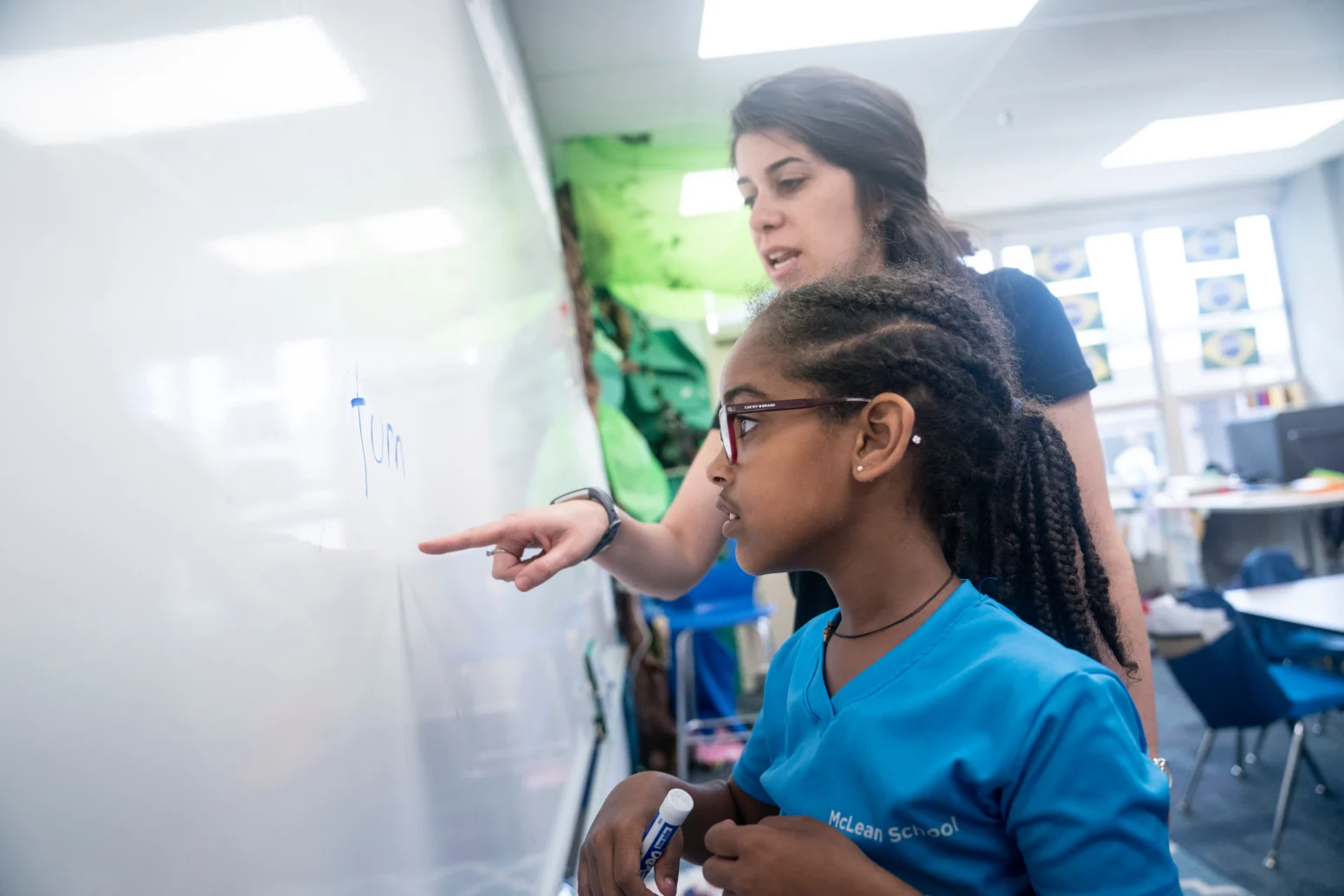 A teacher points at a word on a classroom whiteboard while a student holding a marker listens and looks on attentively.