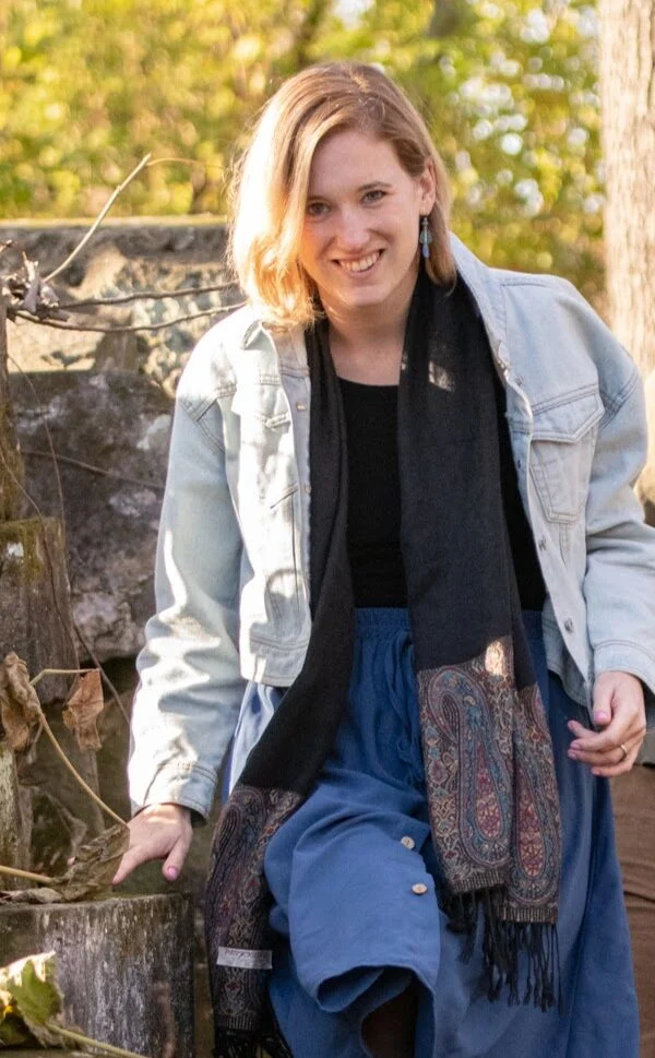 A woman with blonde hair wearing a light denim jacket, black scarf, and blue skirt stands outdoors near stone steps, smiling at the camera.