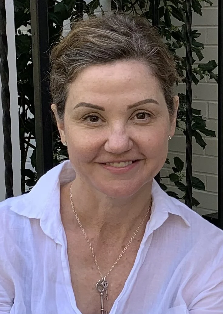 A woman with short brown hair and a white shirt smiles at the camera while sitting outside near a black metal fence and leafy plants.