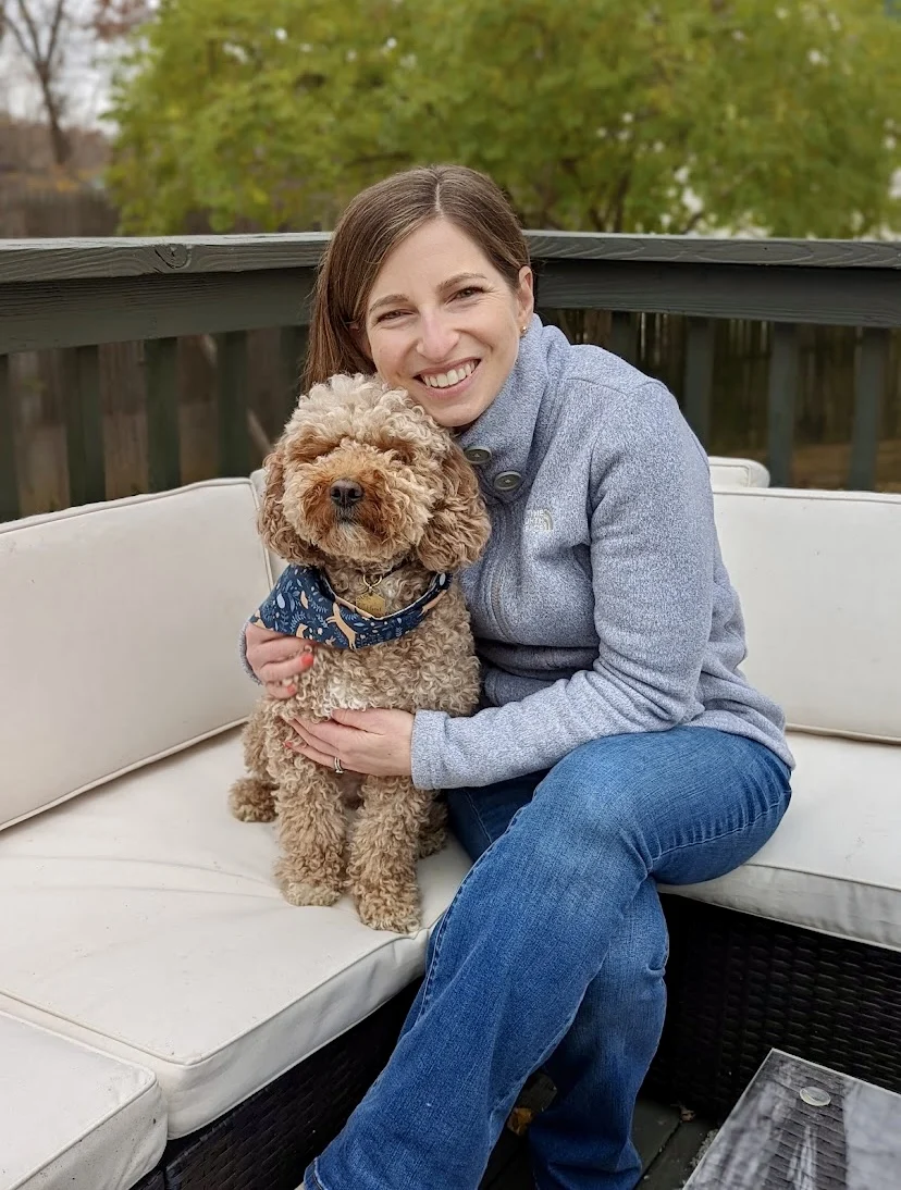 A woman sitting on an outdoor couch smiles while hugging a curly-haired brown dog wearing a blue bandana. Trees and a fence are visible in the background.
