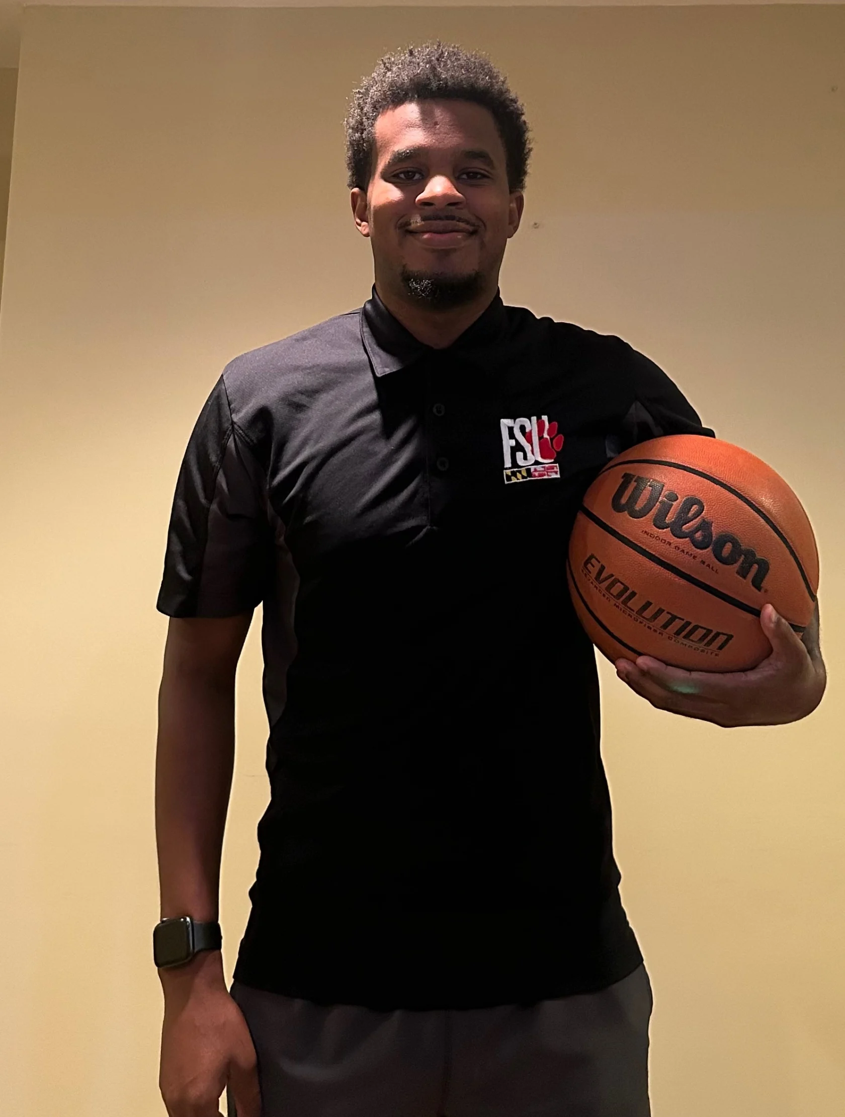 A man wearing a black polo shirt with an "FSL" logo stands indoors holding a Wilson Evolution basketball in his right hand.
