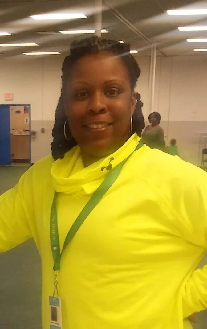 A person wearing a bright yellow top with a lanyard and badge stands indoors, smiling at the camera. Another person is visible in the background.