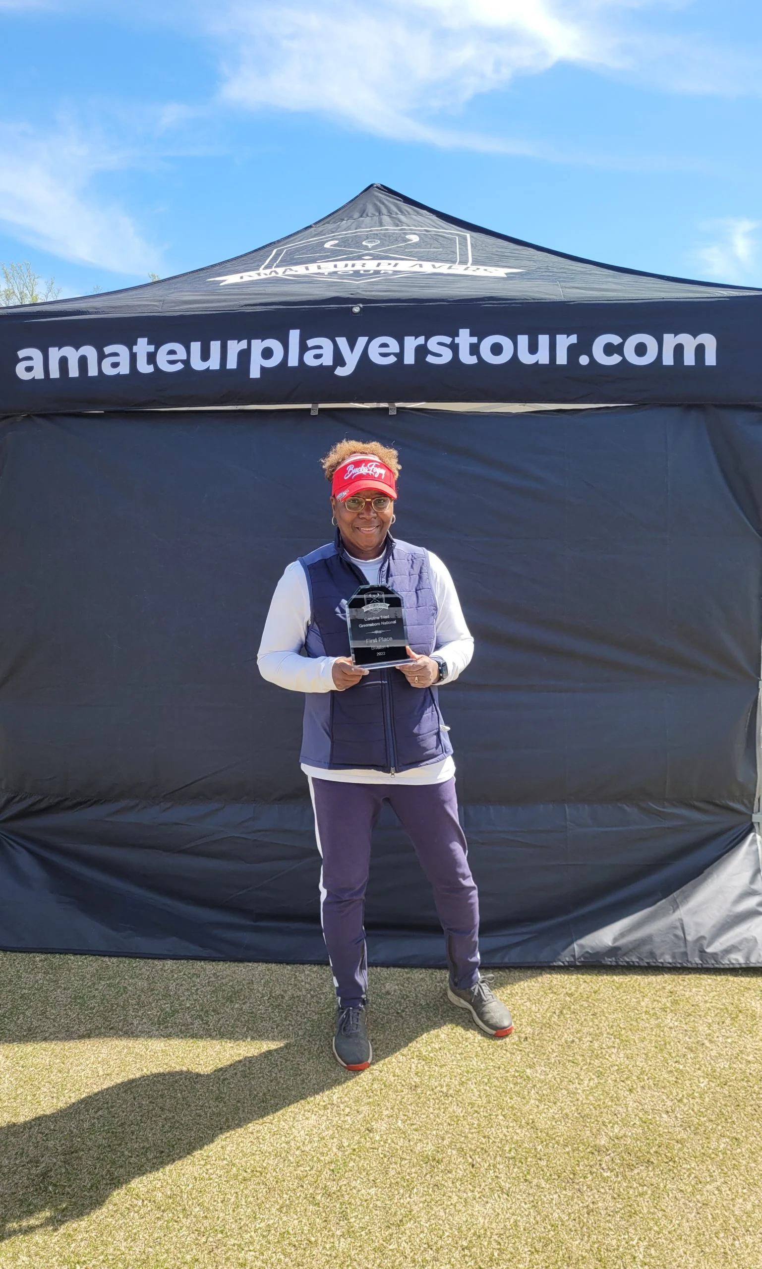 Person standing on grass, holding a trophy in front of a black tent with "amateurplayerstour.com" printed on it, under a blue sky.