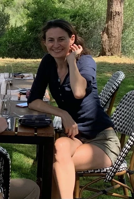 A woman sits outdoors at a table set for a meal, smiling at the camera. She is wearing a navy shirt and shorts, with green trees and grass in the background.